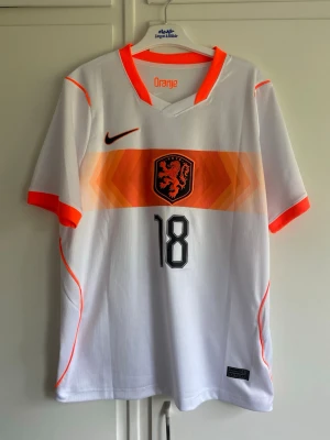 Holland fotbollströja Nike nr 18 - Säljer en vit Holland fotbollströja från Nike med orange detaljer och nummer 18 på både fram- och baksidan. Tröjan har texten 'Oranje' i nacken och 'Malen' på ryggen. Tillverkad i lätt och ventilerande material, perfekt för match eller träning.