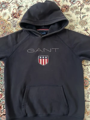 Svart hoodie från GANT med logga - Snygg svart hoodie från GANT med stor broderad logga och sköld med stjärnor och ränder på bröstet. Klassisk känguruficka framtill och huva. Perfekt för dig som gillar stilrena och sportiga plagg. Tillverkad i mjukt material för extra komfort.