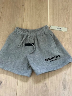 Grå shorts från Essentials Fear of God - Snygga grå shorts från Essentials Fear of God med elastisk midja och snörning. Mjukt material och logga tryckt på ena benet. Perfekta för en avslappnad och trendig look. Kid size