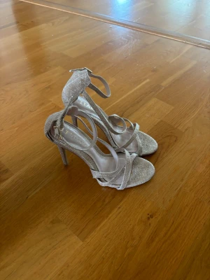 Glittriga silverfärgade pumps - Säljer ett par glittriga pumps i silver med smala remmar och öppen tå. Skorna har hög klack och justerbart ankelband för extra stöd. Perfekta för dig som vill sticka ut och glänsa lite extra. Materialet är glittrigt tyg som fångar ljuset snyggt.