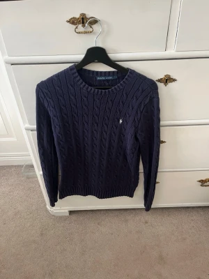 Mörkblå kabelstickad tröja Ralph Lauren - Kabelstickad tröja i mörkblått från Ralph Lauren med rund halsringning och diskret logga broderad på bröstet. Tröjan har långa ärmar och ribbade muddar vid ärmslut och nederkant. Perfekt för lager på lager och klassisk stil.