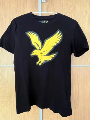 Svart t-shirt med gul örn Lyle & Scott - Snygg svart t-shirt från Lyle & Scott med stor gul örn på bröstet. Klassisk rund hals och korta ärmar. Tillverkad i mjuk bomull som känns skön mot huden. Perfekt för dig som gillar stilrena men ändå iögonfallande plagg.