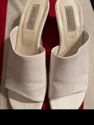 Vita slip-in sandaler med klack - Säljer ett par vita slip-in sandaler från Vander Laan. De har öppen tå, bred rem i syntetläder och en fyrkantig klack i brunt. Perfekta för dig som gillar enkel och stilren design med en liten klack för extra höjd.