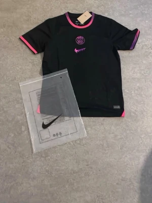 Svart PSG t-shirt med rosa detaljer - Snygg svart t-shirt med Paris Saint-Germain-logga i rosa på bröstet och rosa detaljer vid ärmslut och krage. Perfekt för dig som gillar fotboll och vill ha en sportig look. Materialet är lätt och andas, vilket gör den skön att ha på sig under träning eller match.