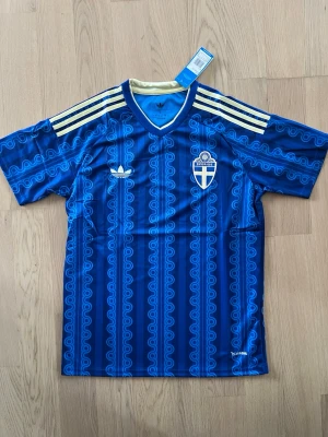 Blå Sverige fotbollströja Adidas - Säljer en blå fotbollströja från Adidas med Sveriges landslagsemblem på bröstet. Tröjan har gula detaljer, klassiska Adidas-ränder på axlarna och ett retroinspirerat mönster. Tillverkad i lätt och ventilerande material, perfekt för träning eller match.
