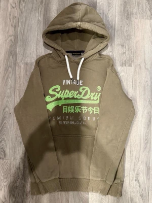 Superdry Hoodie Khaki Strl M - Superdry grön khaki hoodie med klassisk passform som funkar till allt, storlek M, bröstvidd 55 cm, axelbredd 49,5 cm, armlängd 64 cm, midja 45 cm, längd 72 cm, i fint skick.