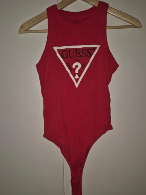 Röd body från Guess med logga - Snygg röd body från Guess med klassisk vit och röd logga framtill. Ärmlös modell med rund halsringning och tryckknappar i grenen. Perfekt att styla till jeans eller kjol för en trendig look.