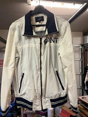  Gant "The Ocean Patch Jacket"  - Snygg vit vindjacka från GANT "The Ocean Patch Jacket" med svarta detaljer på krage, muddar och dragkedjor. Jackan har två fickor med dragkedja och stor GANT-logga på bröstet. Perfekt för blåsiga dagar och enkel att matcha med olika outfits. Svarar gärna om du vill veta storlek i cm, skriv bara vilka mått du vill ha. //
