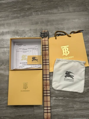 Beige rutigt bälte från Burberry - Säljer ett klassiskt beige bälte från Burberry med det ikoniska rutiga mönstret i brunt, svart och rött. Bältet har ett elegant metallspänne med Burberry-logga och kommer med originalförpackning och dustbag.