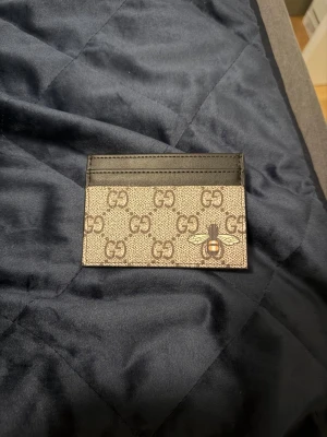 Zara Korthållare  - Snygg korthållare i beige och brunt med klassiskt GG-mönster och en cool bi-detalj på framsidan. Tillverkad i canvas och läder med flera fack för kort. Perfekt accessoar för dig som vill ha något stilrent och lyxigt i fickan.