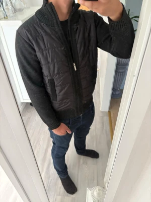 Svart stickad cardigan med quiltade detaljer - Svart cardiganjacka med stickade ärmar och rygg samt quiltad framsida. Jackan har hög krage, dragkedja och två fickor framtill. Insidan har ett zebramönstrat foder som ger en cool kontrast. Perfekt för lager-på-lager-stil. Priser kan ändras vid snabb o smidig affär.