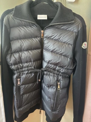 Svart cardigan jacka från Moncler - Snygg svart cardigan jacket från Moncler med quiltad front och stickade ärmar. Jackan har dragkedja framtill, två dragkedjefickor och justerbar midja med snörning. Moncler-logga på ärmen och ribbade muddar vid krage och ärmslut. Perfekt för vår höst sommar 