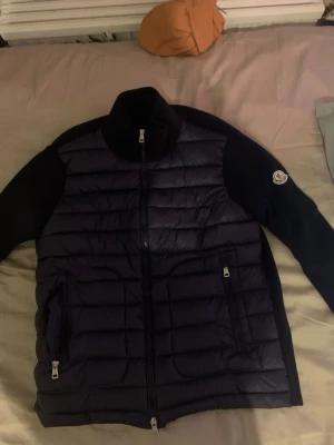 Mörkblå dunväst från Moncler - Snygg mörkblå dunväst från Moncler med stickade ärmar och quiltad front. Västen har dragkedja framtill, två fickor med dragkedja och Moncler-logga på ärmen. Perfekt för lager-på-lager och riktigt trendig look.