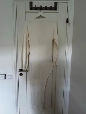 Långärmad vit ribbad långklänning - Säljer en elegant vit långklänning med lång ärm och ribbad struktur. Klänningen har en asymmetrisk nederkant som ger en modern touch. Perfekt för dig som gillar stilrena och minimalistiska plagg.