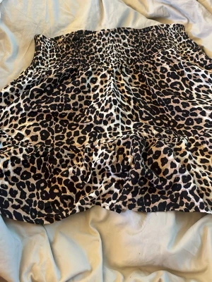 Kort leopardmönstrad kjol - Säljer en kort kjol med leopardmönster i beige, svart och bruna toner. Kjolen har en bred resår i midjan för skön passform och ett lätt, flödande tyg som ger en avslappnad look. Perfekt för dig som vill sticka ut med ett djuriskt print.