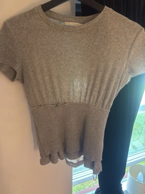Grå ribbad topp med volangkant - Snygg grå ribbad topp med korta ärmar och rund hals. Toppen har en markerad midja och volangkant nedtill som ger en söt och trendig look. Perfekt att matcha med jeans eller kjol för en avslappnad stil.