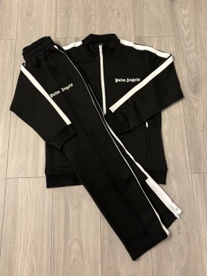 Palm Angels svart tracksuit set - Snyggt svart tracksuit set från Palm Angels med vita ränder längs ärmar och ben. Setet består av en zip-jacka och matchande byxor, båda med logotypen tryckt i vitt. Materialet är mjukt och stretchigt, perfekt för en sportig och avslappnad look.