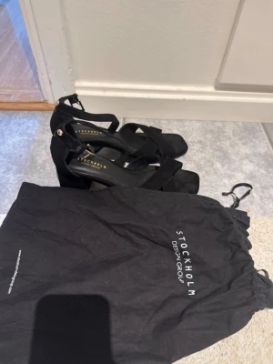 Svarta sandaler från Stockholm Design Group - Snygga svarta sandaler med breda korsade remmar och justerbart spänne runt ankeln. Klackarna är chunky och sulan har en fyrkantig form. Materialet ser ut att vara mockaimitation. Perfekta för dig som vill ha en stilren och trendig look.