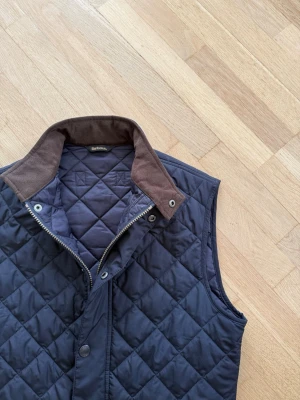 Mörkblå quiltad dunväst Barbour - Snygg mörkblå quiltad dunväst från Barbour med snygga bruna detaljer på kragen. Västen har tryckknappar, dragkedja och två stora fickor framtill. Perfekt för lager-på-lager och ger en mycket klassisk look. Västen är i storlek S och är i bra skick, ett litet håll vid höger sida krage men syns inte om kragen är nedfälld ( se sista bilden )