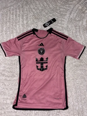 Rosa Inter Miami CF matchtröja Adidas - Säljer en rosa matchtröja från Inter Miami CF med svarta detaljer och klubbmärke på bröstet. Tröjan har svarta ränder på axlarna, svart logga och tryck framtill samt svarta kanter. Perfekt för fotbollsträning eller att bära som supporter.