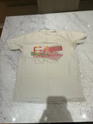 Vit EA7 Emporio Armani t-shirt - Säljer en vit t-shirt från EA7 Emporio Armani med färgglad grafisk logga på bröstet i rött, gult och grönt. Klassisk passform med rund hals och korta ärmar. Perfekt för dig som gillar stilrena plagg med en twist.