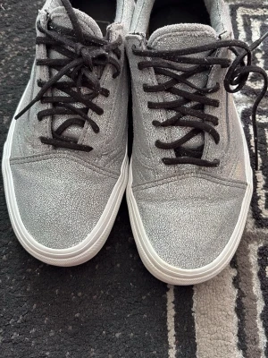 Silvriga Vans sneakers med svarta snören - Snygga silvriga Vans sneakers med glittrig yta och svarta snören. Skorna har en klassisk låg modell med vit sula och svart Vans-logga på innersulan. Perfekta för dig som vill sticka ut med en cool och unik stil.
