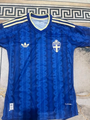 Blå Sverige fotbollströja Adidas - Säljer en blå fotbollströja från Adidas med Sveriges landslagsemblem på bröstet. Tröjan har gula detaljer på axlar och krage, samt ett mönster i tyget. Perfekt för dig som vill stötta Sverige med stil på matchen!