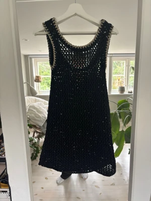 Svart virkad ärmlös klänning - Snygg svart virkad klänning med ärmlös design och rundad halsringning. Klänningen har ett öppet, nätliknande mönster och kontrastkant i beige vid hals och ärmhål. Perfekt att styla över andra plagg för en trendig look.