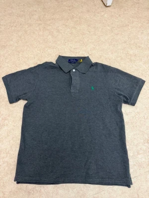 Mörkgrå pikétröja från Polo Ralph Lauren - Klassisk mörkgrå pikétröja från Polo Ralph Lauren med korta ärmar och krage. Tröjan har två knappar vid halsen och den ikoniska gröna broderade loggan på bröstet. Perfekt till jeans eller shorts för en clean look.