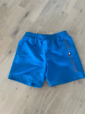 Blå badbyxor från Tommy Hilfiger, stl 12-14y - Snygga blå badbyxor från Tommy Hilfiger med resår och dragsko i midjan. Märkets logga och flagga syns längs ena benet. Bakficka med knapp och liten logodetalj. Perfekta för stranden eller poolen.