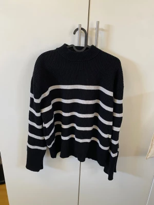 Svartvit randig stickad tröja - Snygg stickad tröja i svart med vita horisontella ränder. Tröjan har en högre ribbad krage och långa ärmar. Perfekt för dig som gillar en klassisk och avslappnad stil. Materialet är grovstickat och ger en mysig känsla.