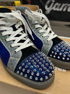 Christian Louboutin sneakers  - Snygga sneakers från Christian Louboutin med blå sammet, silverglittriga detaljer och vita skosnören. Tåpartiet är täckt av silvriga nitar och baksidan har en gul detalj. Skorna har en grå sula och sticker ut med sin unika design.
