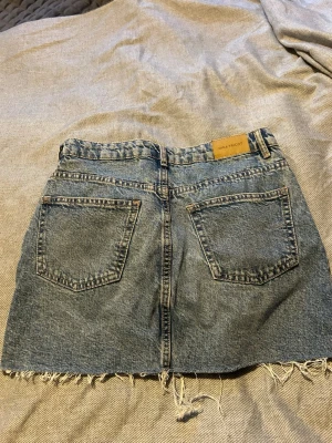 Blå jeanskjol från Gina Tricot - Säljer en klassisk blå jeanskjol från Gina Tricot med rå kant nertill och två bakfickor. Kjolen har en avslappnad passform och är tillverkad i slitstarkt denimtyg. Perfekt för dig som gillar en enkel men snygg stil.