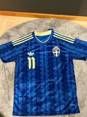 Blå Sverige fotbollströja Adidas - Säljer en blå fotbollströja från Adidas med Sveriges landslagsemblem, gula detaljer och nummer 11. Tröjan har Elanga tryckt på ryggen, korta ärmar och klassiska Adidas-ränder på axlarna. Materialet är lätt och andas, perfekt för träning eller match.