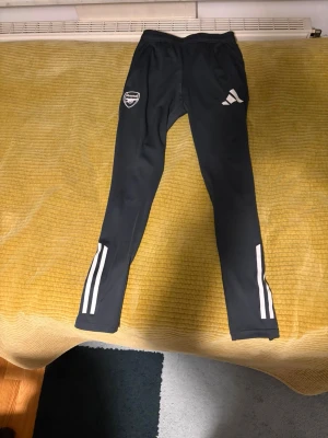Svarta Arsenal träningsbyxor Adidas - Säljer ett par svarta träningsbyxor från Adidas med Arsenal-logga. Byxorna har vita ränder och logotyper på framsidan samt röda detaljer på baksidan. Tillverkade i ett stretchigt funktionsmaterial med smal passform och dragkedjor vid bensluten. Priset kan diskuteras.