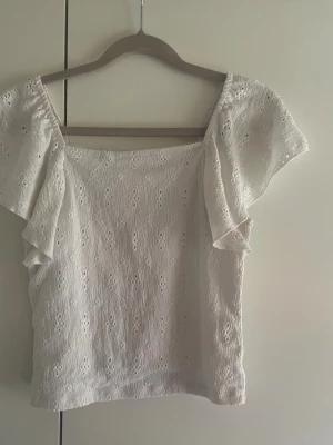 Vit blus med broderade detaljer Kappahl - Söt vit blus från Kappahl med broderade hålmönster och korta, fladdriga ärmar. Blusen har en rak passform och är tillverkad i ett lätt bomullstyg, perfekt för varma dagar. Supersnygg till både jeans och kjol!