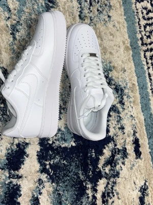 Nike Air Force 1 vita sneakers Helt nya -  Klassiska helvita Nike Air Force 1 sneakers i läder med perforerad tåbox, tjock sula och ikonisk Swoosh på sidan. Skorna har vita skosnören och en stilren, tidlös design som passar till det mesta. Perfekta för dig som gillar streetwear och enkel stil.