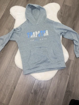 Grå hoodie från Trapstar - Snygg grå hoodie från Trapstar med tryck i blått och vitt på bröstet. Klassisk känguruficka framtill och huva. Perfekt för en avslappnad streetwear-look.