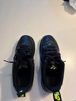 Nike Air Force 1 svart/neongul - Säljer ett par svarta Nike Air Force 1 sneakers med neongula detaljer på loggan och hälen. Skorna är låga och har klassisk siluett med snörning och perforerad tå. Tillverkade i läder med textildetaljer för extra komfort.