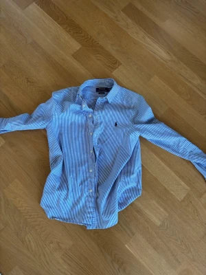 Blåvit randig skjorta Polo Ralph Lauren - Snygg blåvit randig skjorta från Polo Ralph Lauren i slim fit-modell. Skjortan har långa ärmar, klassisk krage och knäppning framtill. Tillverkad i bomull och prydd med den ikoniska loggan på bröstet. Perfekt för en stilren look. Skulle säga att den passar en person som är 13-14 beror på längd. Passade mig när jag var 160-168 och var 13-14 år gammal. 