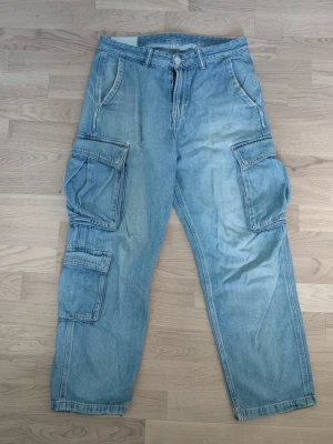 Ljusblå denim cargobyxor - Säljer ett par ljusblå cargobyxor i denim med flera stora fickor på sidorna. Byxorna har avslappnad passform, klassisk knappgylf och bälteshällor. Perfekta för en streetwear-look och tillverkade i bomull. De har en liten lagning vid sömmen vid den ena fickan vilket syns på bild 4. Utöver det är byxorna i gott skick. 