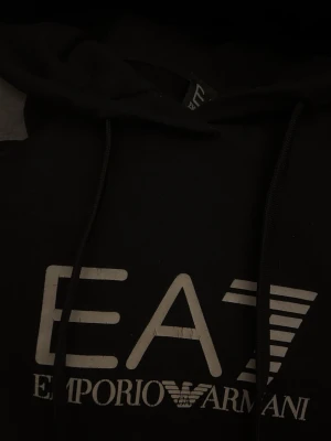 Svart hoodie från Emporio Armani EA7 - Svart hoodie från Emporio Armani EA7 med stor vit logga på bröstet. Klassisk modell med huva och dragsnören. Perfekt för en stilren och sportig look. Mjuk bomullskänsla och avslappnad passform.