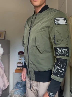 Olivgrön tiger of Sweden ”The Weeknd” bomberjacka med patches - Snygg olivgrön bomberjacka med svarta muddar och coola patches på båda ärmarna. Jackan har dragkedja framtill, två stora fickor med lock och en klassisk bomberkrage. Perfekt för dig som vill sticka ut med detaljer och streetstyle. Jackan kommer från Tiger of Sweden och har väldigt sällan använts. (Passform: jag är 190 lång och väger 80kg)
