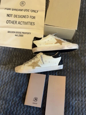 Golden Goose sneakers med glitterdetaljer - Snygga vita Golden Goose sneakers med glittrig guldstjärna på sidan och svartglittrig häl. Skorna har grå skosnören och klassisk låg modell. Perfekta för dig som vill sticka ut med en unik och trendig look.