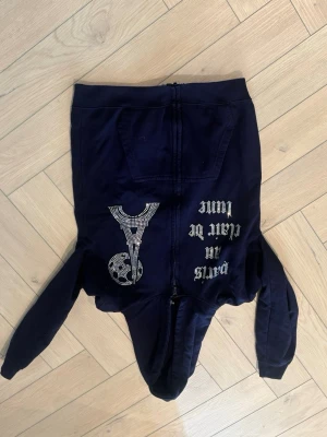 Mörkblå hoodie med tryck och dragkedja - Snygg mörkblå hoodie med dragkedja framtill och huva. På framsidan finns ett coolt tryck med texten 'Paris in clear be lame' och ett glittrigt Eiffeltorn i strasstenar. Hoodien har långa ärmar och känguruficka. Perfekt för en trendig streetwear-look.