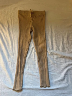 Beige ribbade leggings från H&M - Säljer ett par beige ribbade leggings från H&M i storlek 122. De har en mjuk och stretchig passform med resår i midjan och vågiga kanter vid bensluten. Perfekta för en avslappnad och trendig look.
