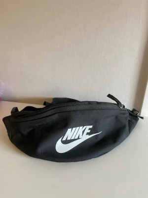 Svart midjeväska från Nike - Snygg svart midjeväska från Nike med klassisk vit logga framtill. Väskan har justerbart band och dragkedja upptill. Tillverkad i slitstarkt tygmaterial och perfekt för att bära det viktigaste. Enkel och sportig design som funkar till många stilar.