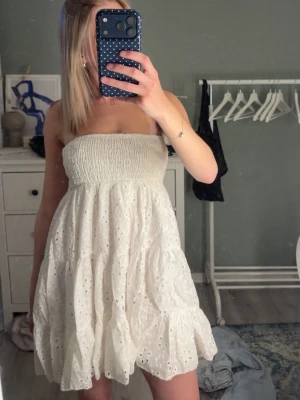 Vit axelbandslös kort klänning - Söt vit axelbandslös klänning med smock upptill och broderade hål i tyget. Köpte förra året och endast använd 1 gång❤️