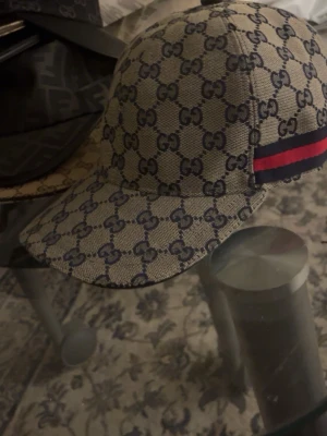 Gucci Navy keps med GG-mönster - Säljer en Navy keps från Gucci med klassiskt GG-monogram i mörkblått över hela kepsen. På sidan finns en ikonisk röd och marinblå rand. Kepsen har böjd skärm och är tillverkad i canvasmaterial. Perfekt för dig som gillar exklusiva accessoarer.
