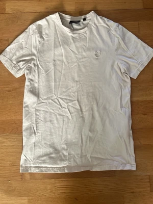 Beige t-shirt från Lyle & Scott - Klassisk beige t-shirt från Lyle & Scott med rund halsringning och diskret broderad logga på bröstet. T-shirten är i 100% bomull och har en enkel, stilren design som passar till allt.
